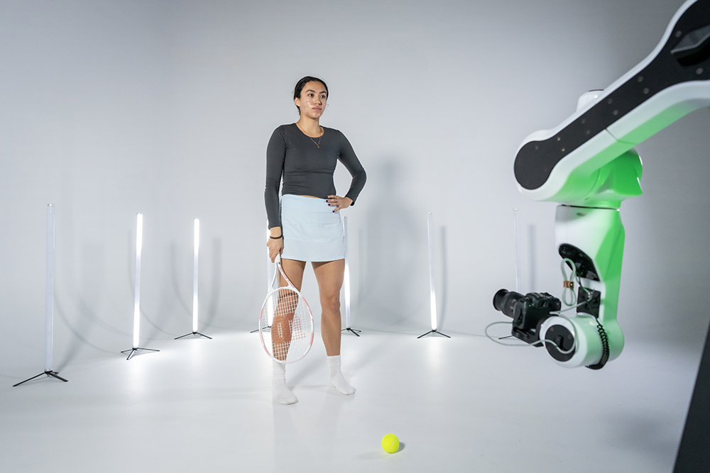 A person with a tennis racket poses in a studio with a tennis ball on the floor, illuminated by vertical lights and facing a robotic camera arm.