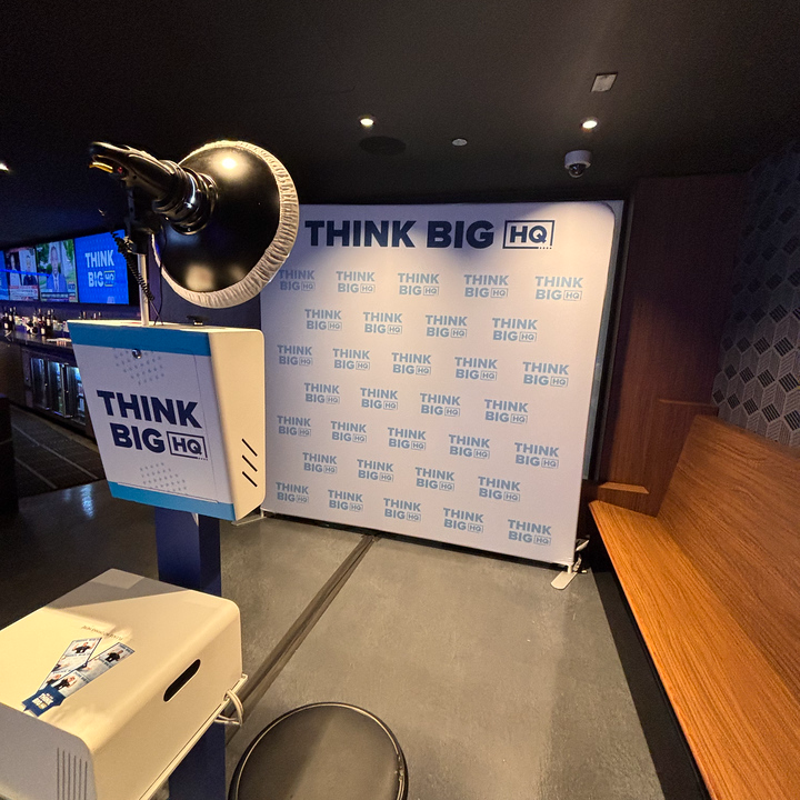 Experience the DNC Photo Booth Experience with a sleek setup featuring a ring light and THINK BIG HQ banner. The modern design includes a wooden bench and patterned wall, creating a bright and professional ambiance perfect for memorable snapshots.