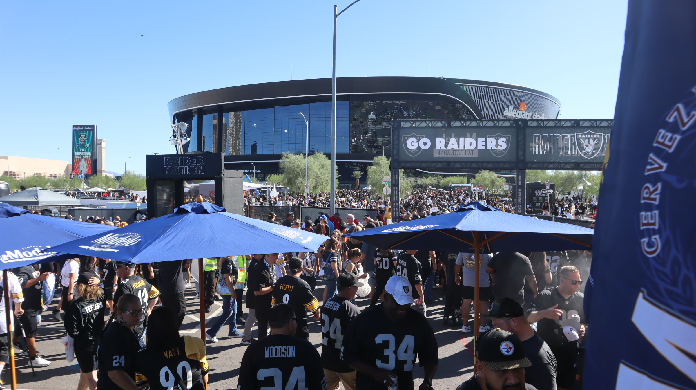 Crowd outside a large stadium, many wearing team jerseys labeled 34 and Watt. Go Raiders signs are visible. Blue umbrellas provide shade. A nearby banner reads Raider Nation. The stadium is Allegiant Stadium, and its a sunny day.