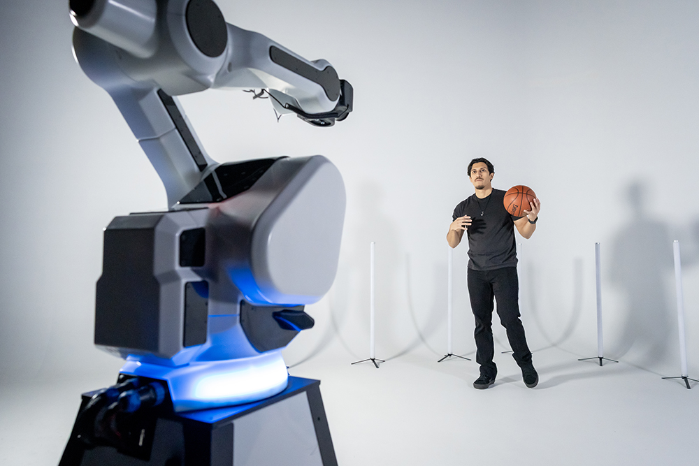 A man holding a basketball stands in front of a robotic arm in a white room, reminiscent of the high-tech setups at photo booth rentals in Chicago.