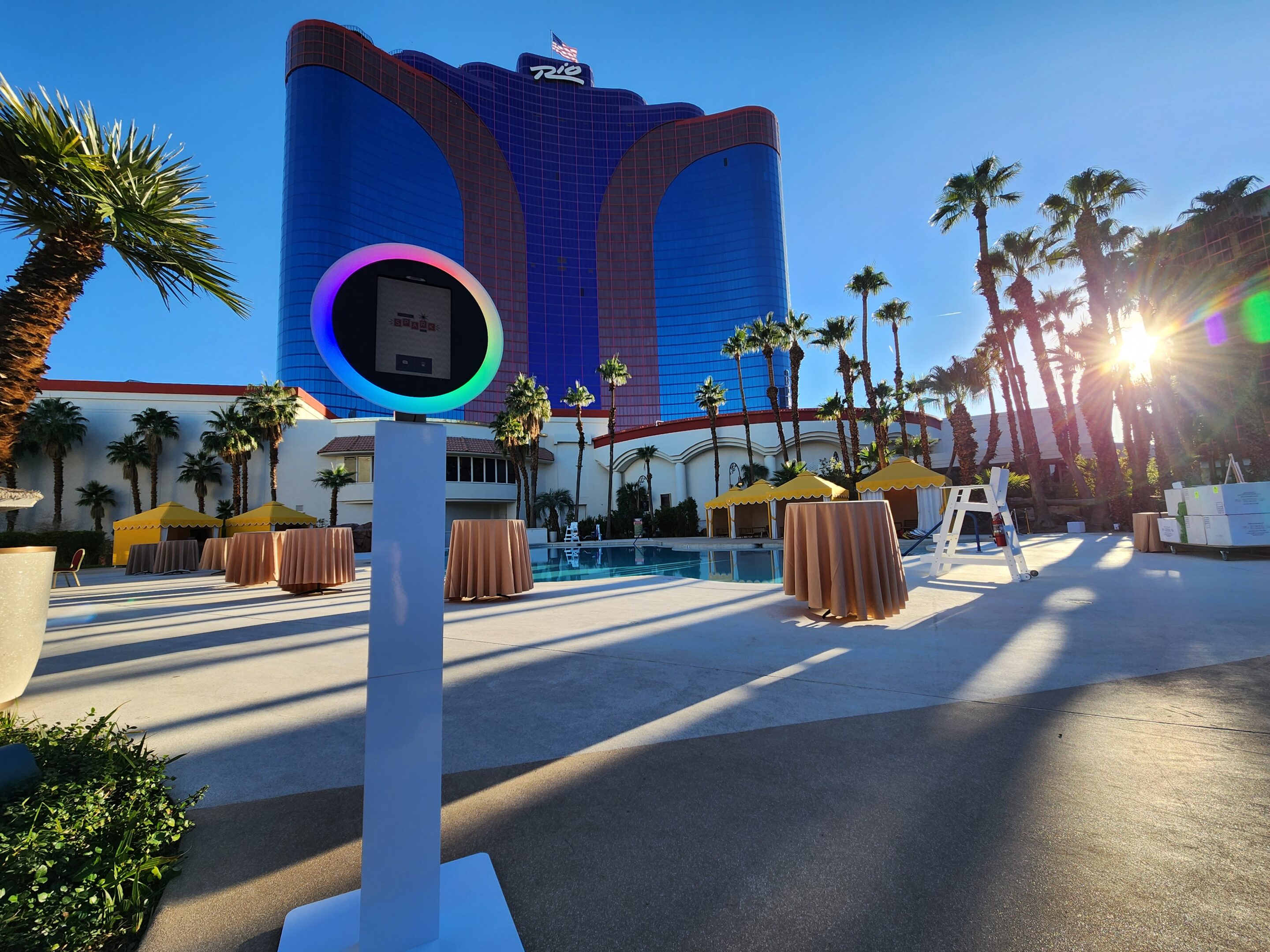 A poolside event setup with cocktail tables and a Las Vegas Photo Booth Rental in front of a tall blue and purple hotel, palm trees, and bright sunlight in the background.