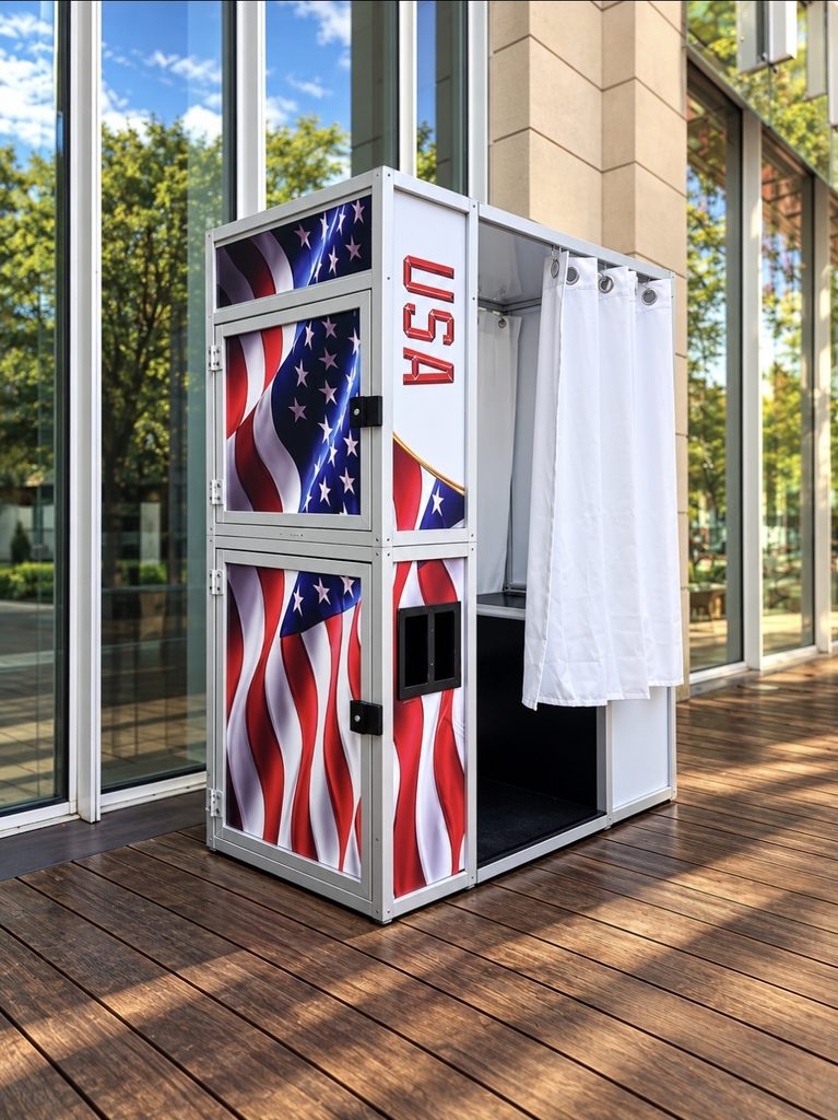 A small experiential photo booth decorated with USA flags and a white curtain stands on a wooden deck outside a glass building.